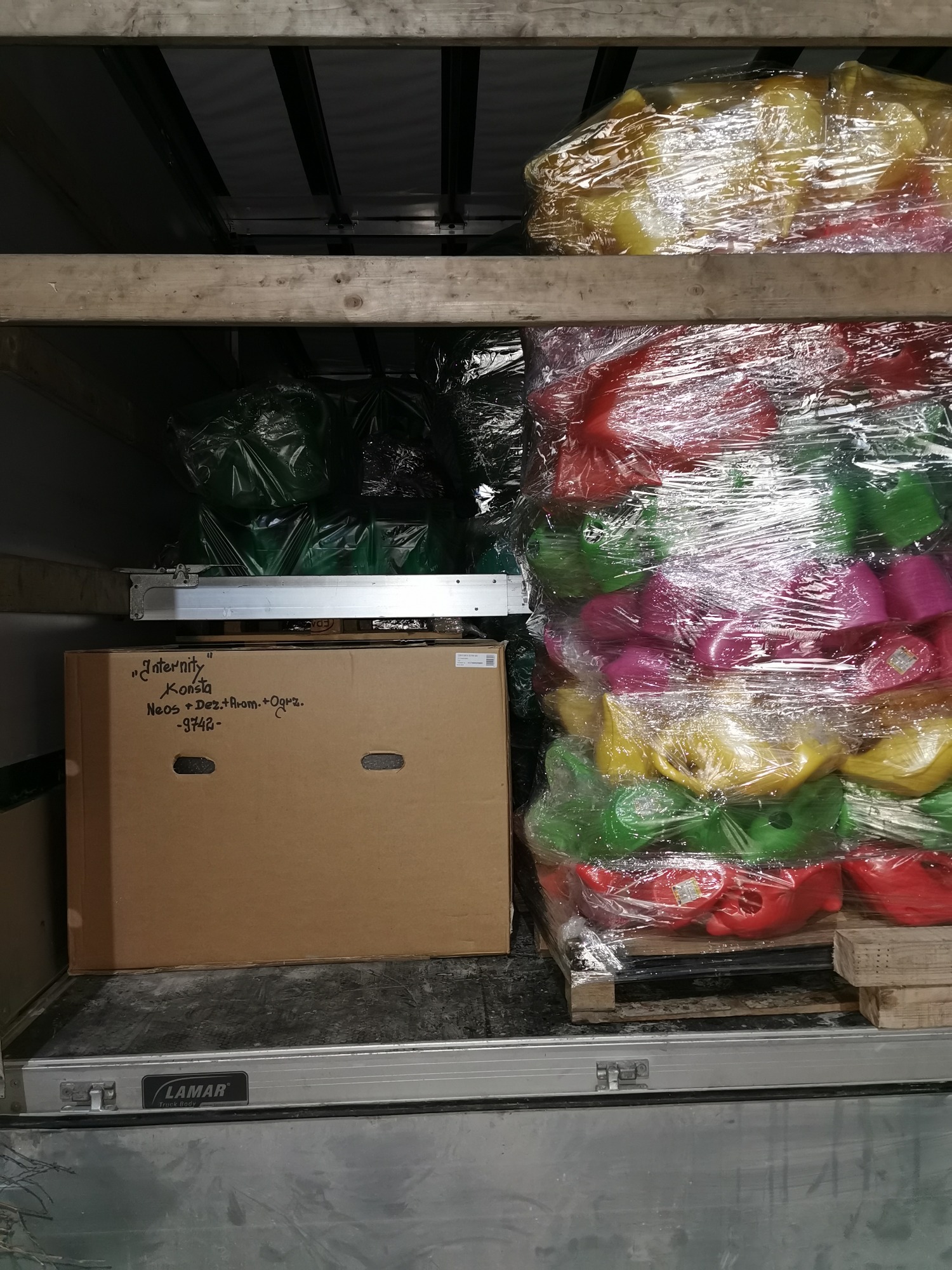 Wnętrze przestrzeni ładunkowej busa: palety z zapakowanymi w folię stretch kolorowymi plastikowymi zabawkami (czerwone, zielone, żółte, różowe) oraz karton z odręcznym napisem 'Internity Konsta...