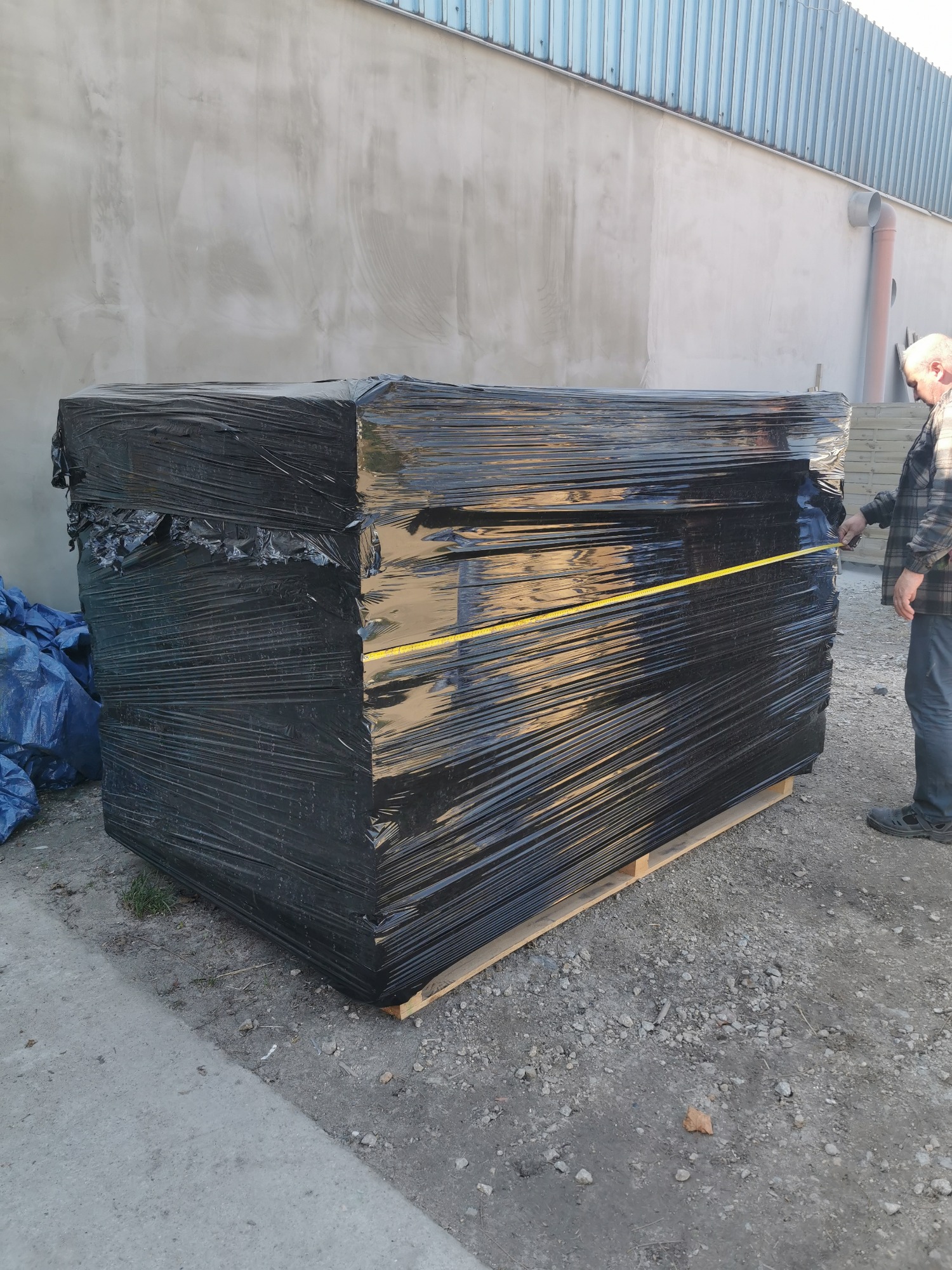 Paczka czarnych płyt zabezpieczonych folią stretch na palecie, mierzona żółtą taśmą mierniczą przez mężczyznę w kraciastej koszuli, na zewnątrz budynku.