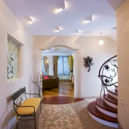 Przestronny hol z dekoracyjnym oświetleniem sufitowym, mozaikową podłogą i schodami z metalową balustradą, widok na salon z okrągłą sofą i stylowymi zasłonami.