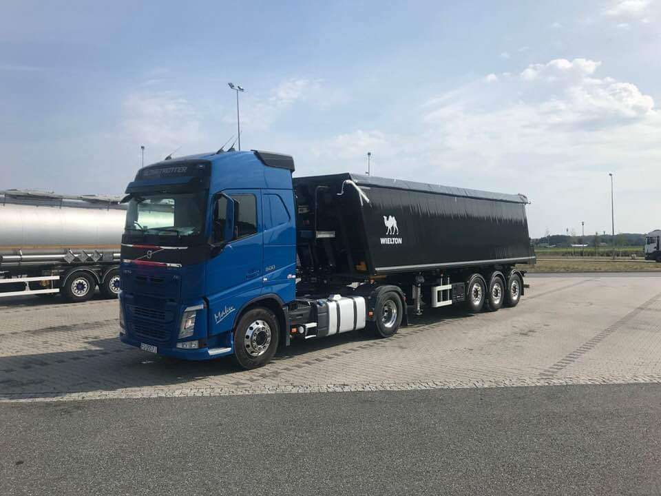 Niebieski ciągnik siodłowy Volvo z czarną naczepą wywrotką Wielton, stojący na parkingu z kostki brukowej w słoneczny dzień.