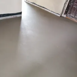 Świeżo wylana, gładka, szara posadzka w pomieszczeniu, z widocznymi taśmami dylatacyjnymi przy ścianach i starą, drewnianą stolarką drzwiową w tle.