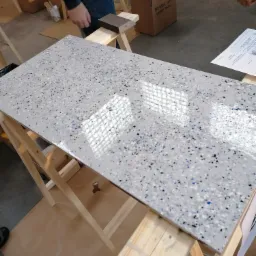 Błyszcząca, szara powierzchnia z kolorowymi drobinkami (niebieskie, czarne, szare) na drewnianym stelażu, imitująca wykończenie podłogi, z widocznymi elementami otoczenia warsztatowego.