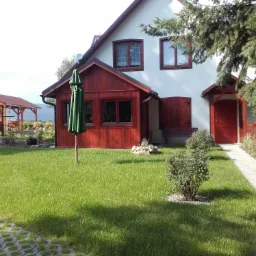 Widok na zadbany trawnik przed domem z drewnianą werandą, widoczny również fragment chodnika z ażurowych płyt i altana w tle.