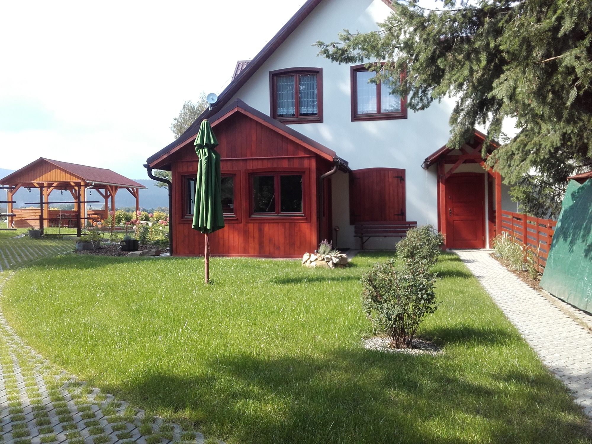 Widok na zadbany trawnik przed domem z drewnianą werandą, widoczny również fragment chodnika z ażurowych płyt i altana w tle.