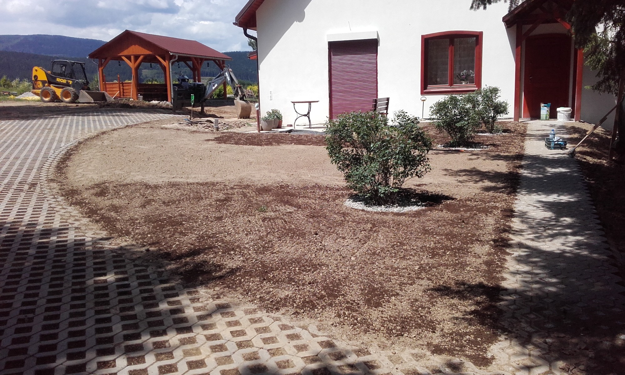 Przygotowanie terenu pod trawnik: wyrównana ziemia, obrzeża z ażurowych płyt betonowych, w tle budynek z czerwoną stolarką i maszyny budowlane.