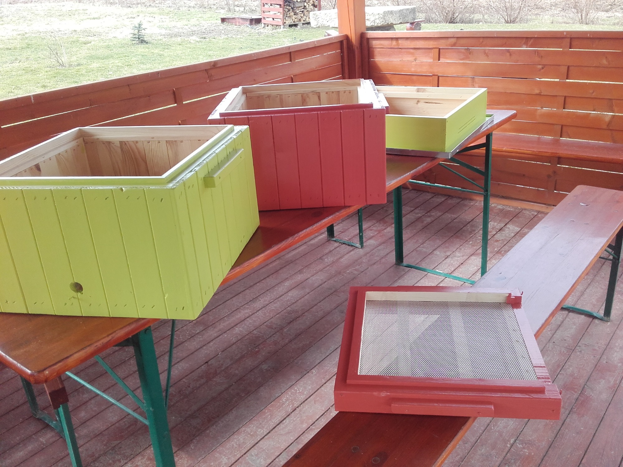 Drewniane, kolorowe skrzynki (żółte, czerwone) na stole w altanie, prawdopodobnie ule pszczele lub elementy uli, z widoczną siatką w jednej z ramek.