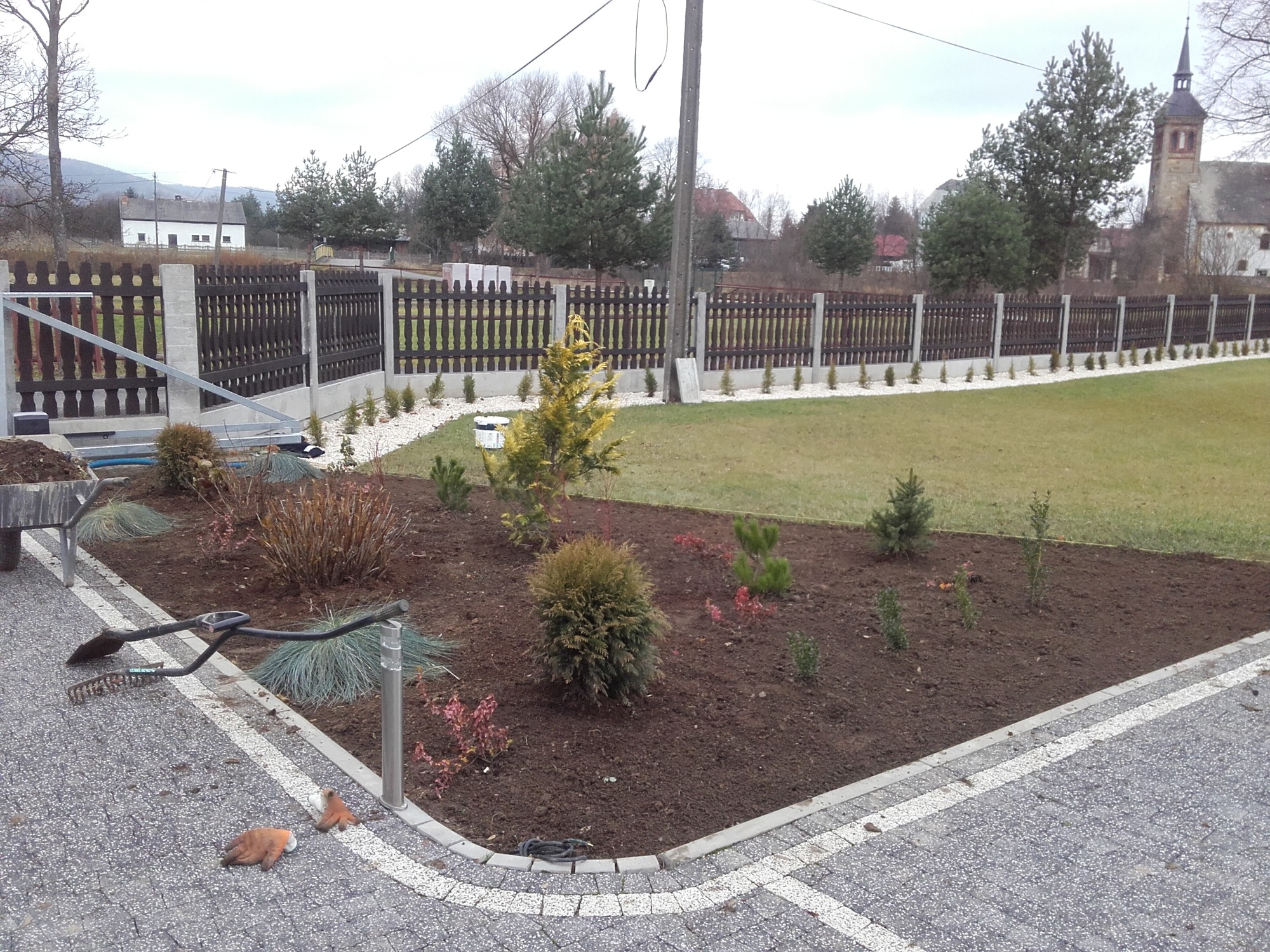 Świeżo założony rabat z różnorodnymi krzewami i drzewkami iglastymi, obok leżą narzędzia ogrodnicze i rękawice, w tle drewniany płot z betonowymi słupkami i kościół.