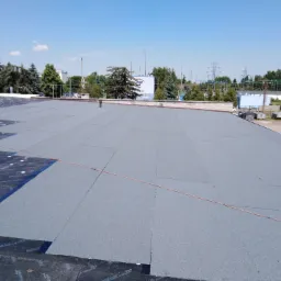 Widok z góry na płaski dach pokryty papą termozgrzewalną, częściowo ułożoną na warstwie izolacyjnej z widocznymi otworami, z butlą gazową w rogu i elementami infrastruktury miejskiej w tle.