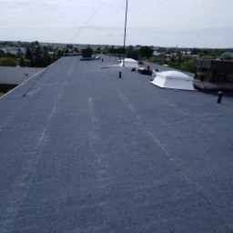 Płaski dach pokryty papą termozgrzewalną z widocznymi świetlikami dachowymi, anteną i elementami wentylacji na tle zabudowy miejskiej.