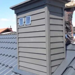 Szary komin obudowany panelami z dwoma kratkami wentylacyjnymi, zamontowany na metalowym dachu, widziany z dołu, z pracownikiem w tle.