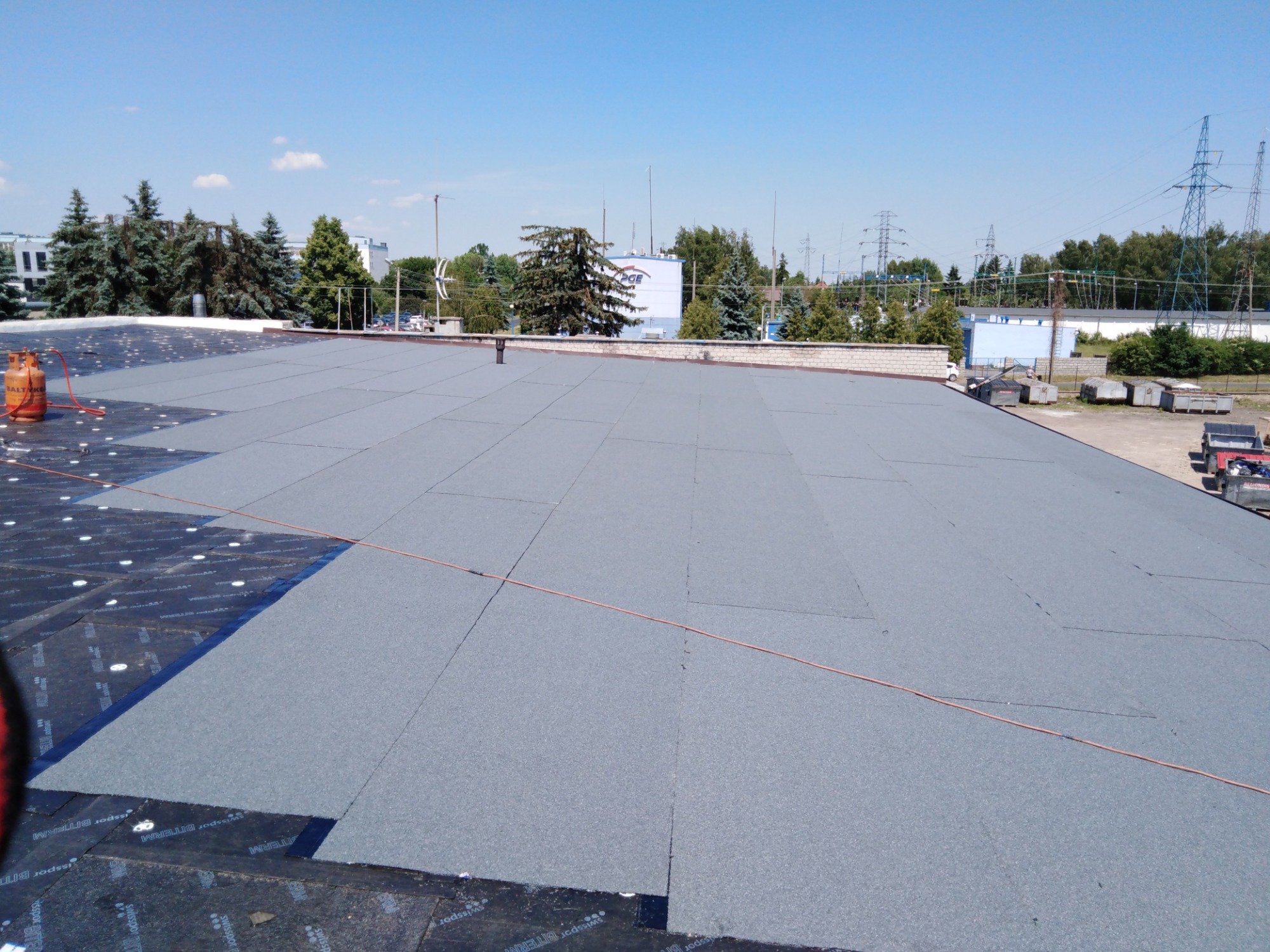 Widok z góry na płaski dach pokryty papą termozgrzewalną, częściowo ułożoną na warstwie izolacyjnej z widocznymi otworami, z butlą gazową w rogu i elementami infrastruktury miejskiej w tle.