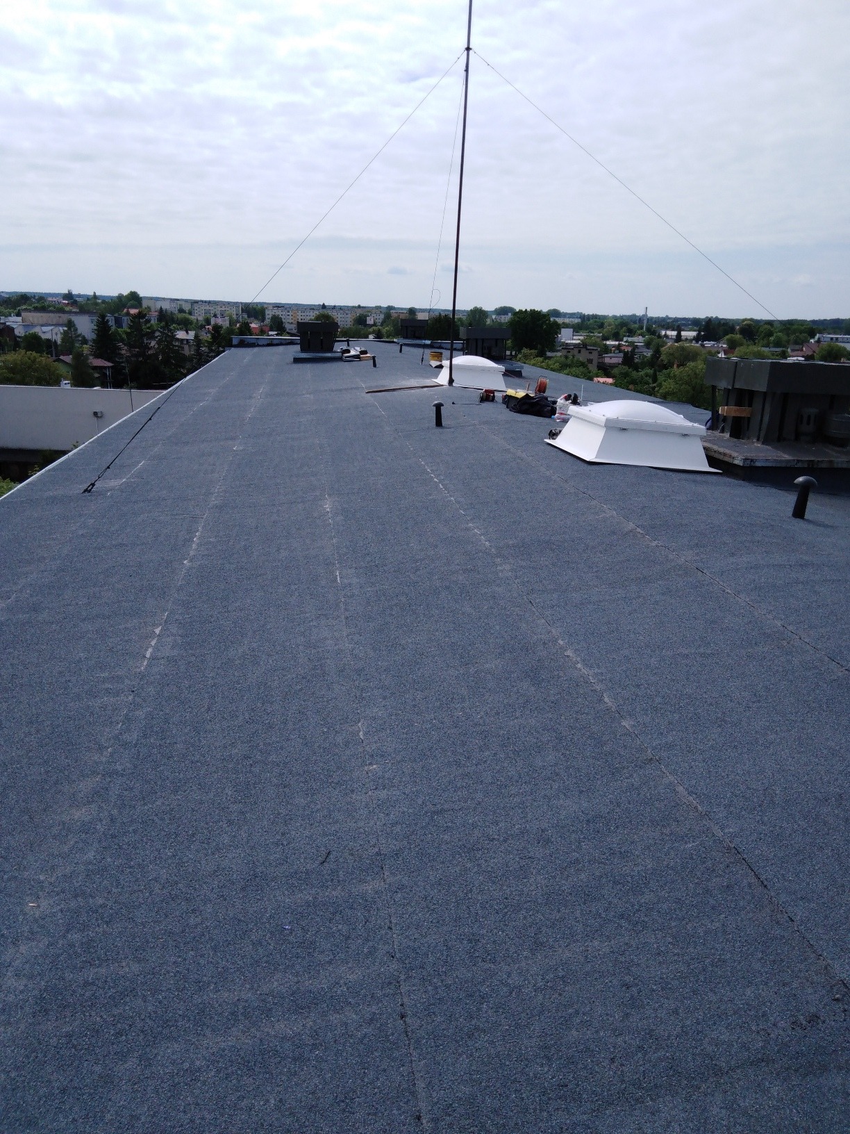 Płaski dach pokryty papą termozgrzewalną z widocznymi świetlikami dachowymi, anteną i elementami wentylacji na tle zabudowy miejskiej.