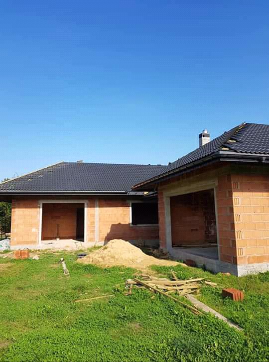 Budynek w trakcie budowy z widocznym czarnym dachem z dachówki ceramicznej i kominem, ściany z czerwonej cegły, na trawniku leżą drewniane elementy.