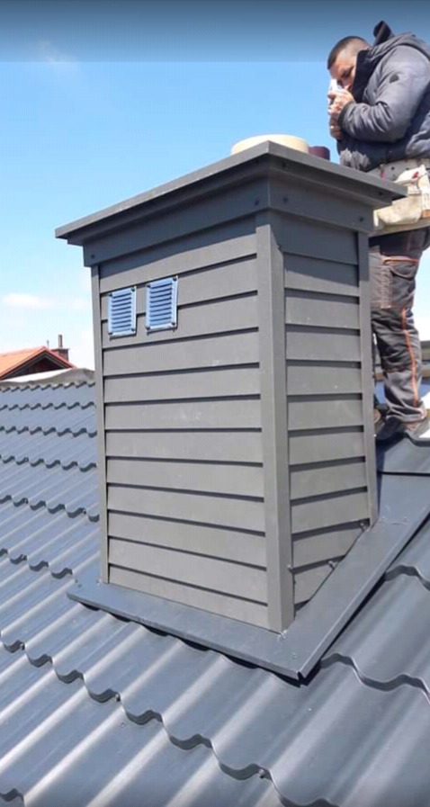 Szary komin obudowany panelami z dwoma kratkami wentylacyjnymi, zamontowany na metalowym dachu, widziany z dołu, z pracownikiem w tle.