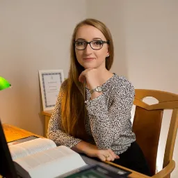 Portret kobiety w okularach siedzącej przy biurku z otwartą księgą, zieloną lampką i certyfikatem w tle.