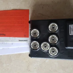 Czarny moduł sterujący Jungheinrich z sześcioma okrągłymi złączami, etykietą z numerem seryjnym i instrukcjami w różnych językach, leżący na beżowym tle.