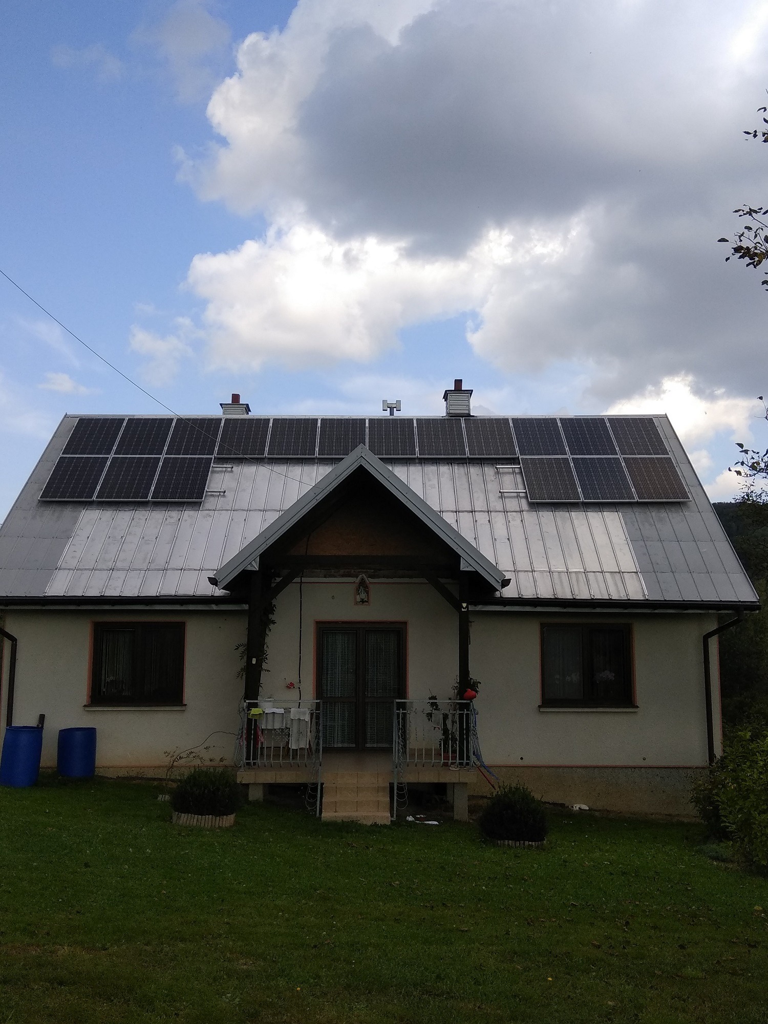 Dom jednorodzinny w Olszynach z panelami słonecznymi na srebrnym dachu, widoczne dwa niebieskie pojemniki na trawniku.