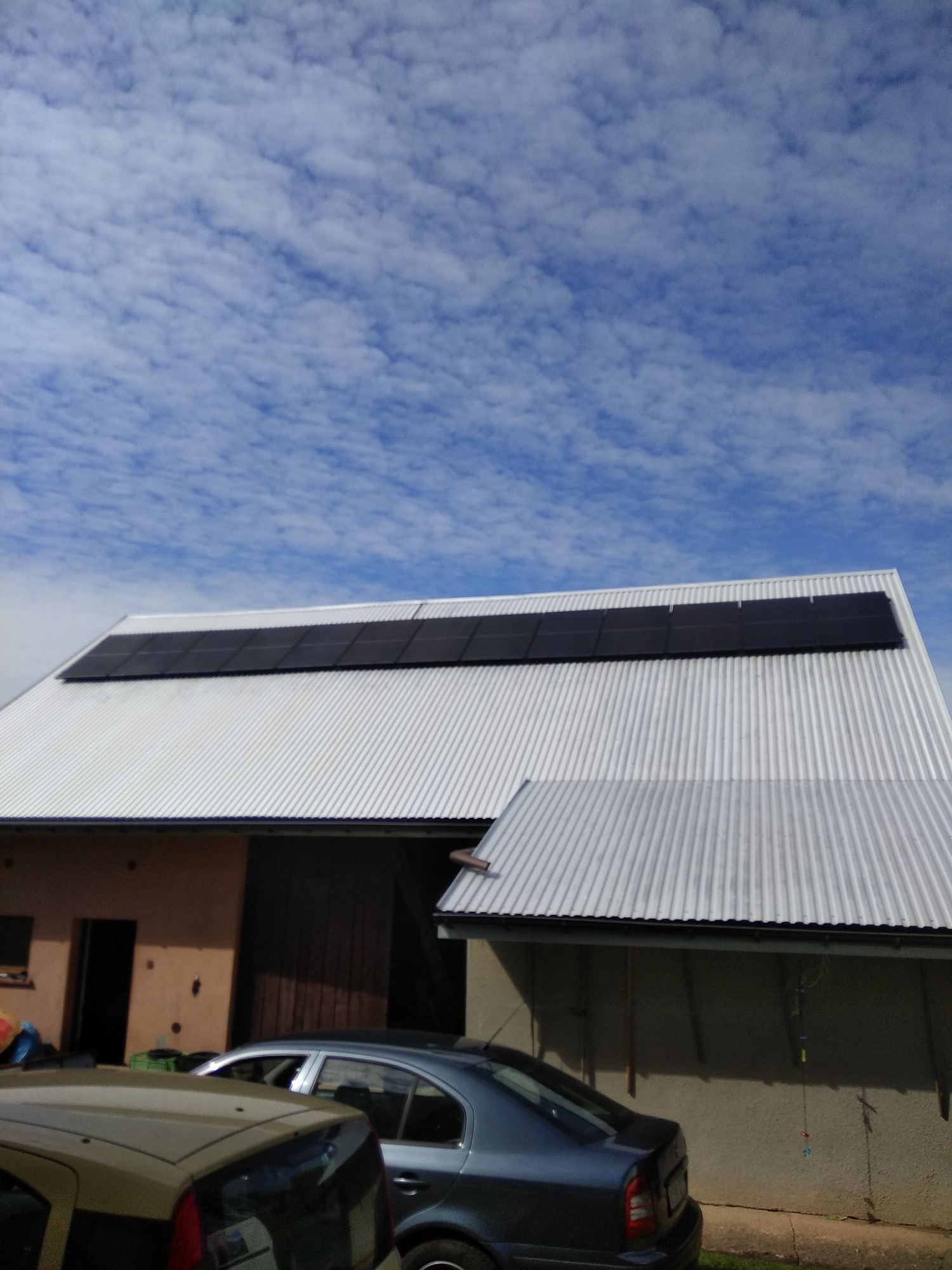 Panel fotowoltaiczny zamontowany na falistym, metalowym dachu budynku gospodarczego, widoczne dwa samochody w tle, błękitne niebo z chmurami.