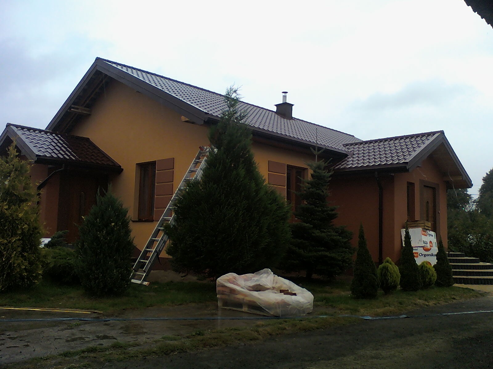 Dom jednorodzinny z nowym, brązowym dachem z blachodachówki, drabina oparta o ścianę, paleta z materiałami budowlanymi zabezpieczona folią, widoczne elementy ogrodu.