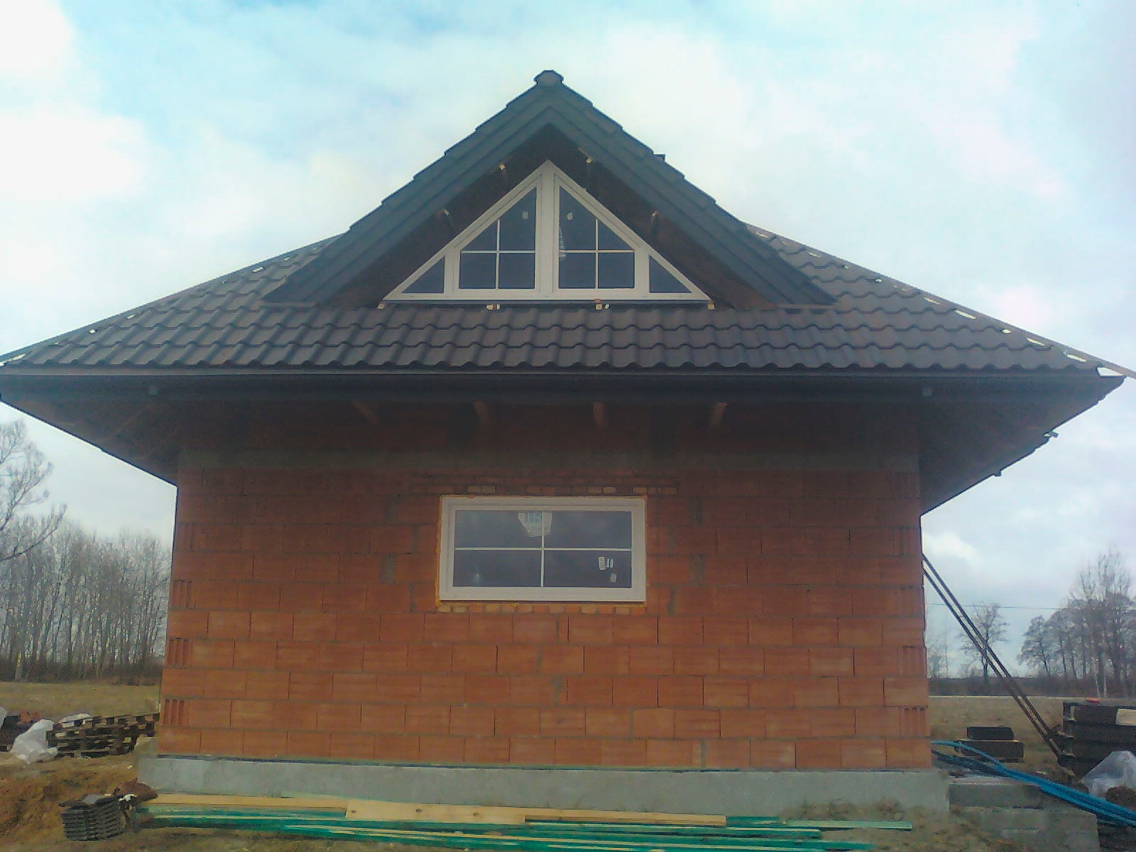 Niedokończony budynek z brązową dachówką ceramiczną, widoczne okno trójkątne na poddaszu i prostokątne na parterze, ściany z czerwonej cegły, w tle drzewa bez liści.