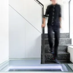 Wnętrze domu z nowoczesnymi schodami z ciemnego metalu z perforowanym wzorem, szklana podłoga z podświetleniem LED, białe ściany i osoba schodząca po schodach.