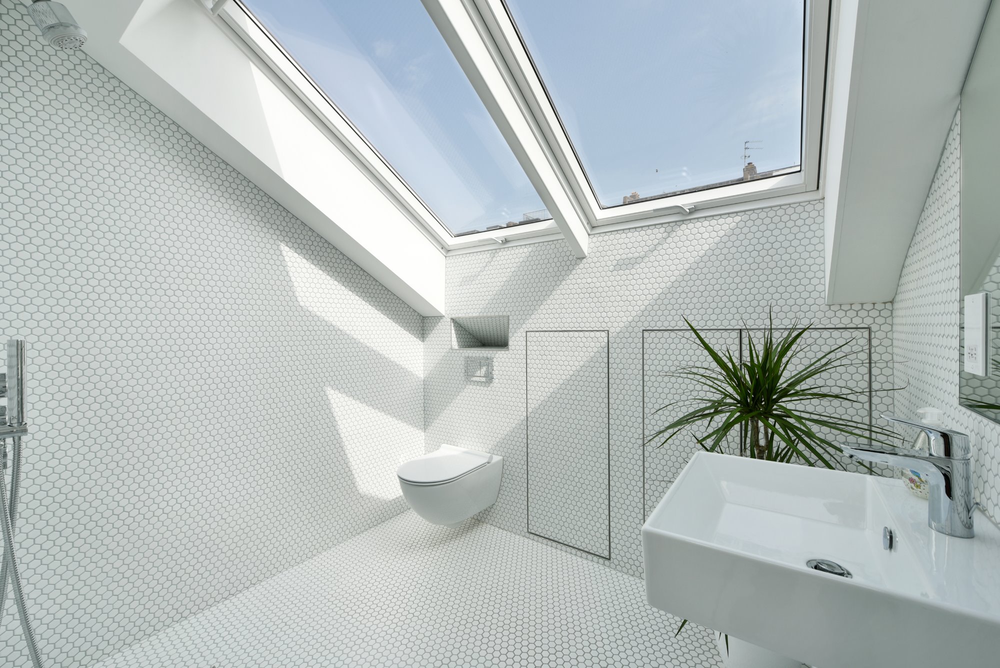 Jasna łazienka na poddaszu z białymi, heksagonalnymi płytkami, oświetlona przez okna dachowe; widoczna toaleta podwieszana, minimalistyczna umywalka i roślina doniczkowa.