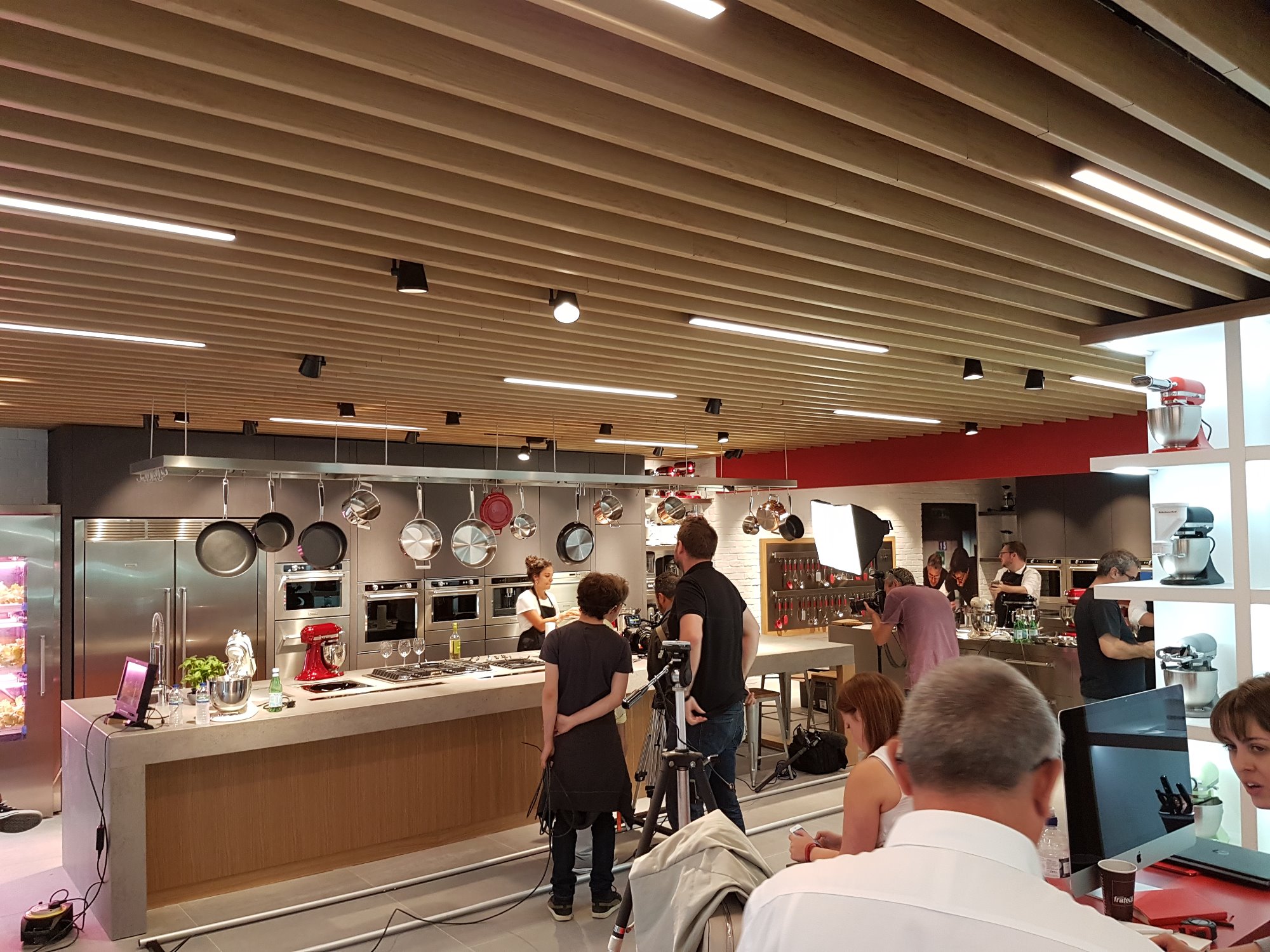 Ekipa filmowa kręci ujęcia w przestronnej kuchni pokazowej z wyspą, wiszącymi garnkami i nowoczesnym sprzętem AGD.
