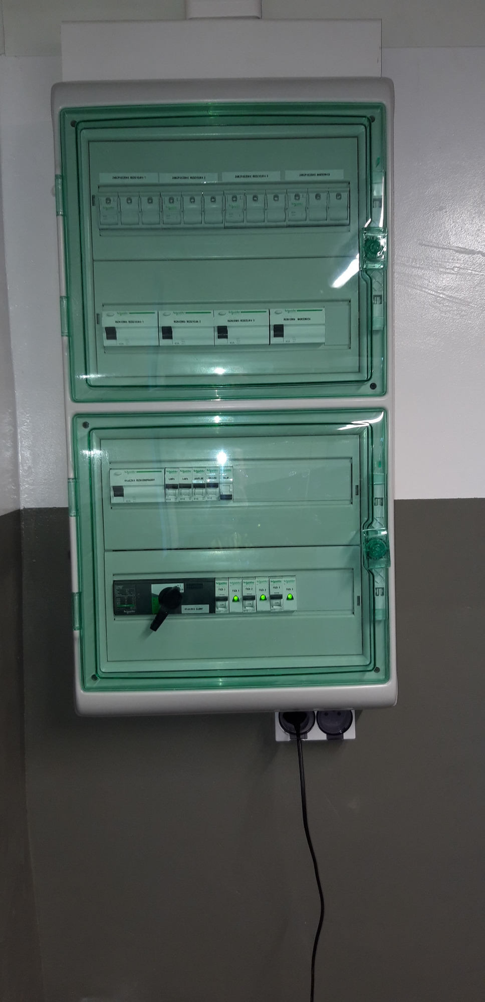 Dwu-segmentowa, zielonkawa skrzynka rozdzielcza Schneider Electric z widocznymi bezpiecznikami i włącznikiem głównym w pozycji 'wyłączony', zamontowana na szaro-białej ścianie, z podłączonym...