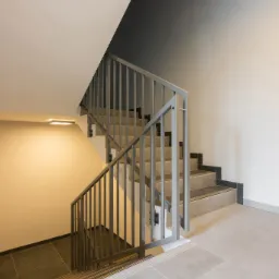 Klatka schodowa z szarymi płytkami podłogowymi i schodami, metalowa balustrada, minimalistyczne oświetlenie sufitowe, widok z góry na dół.