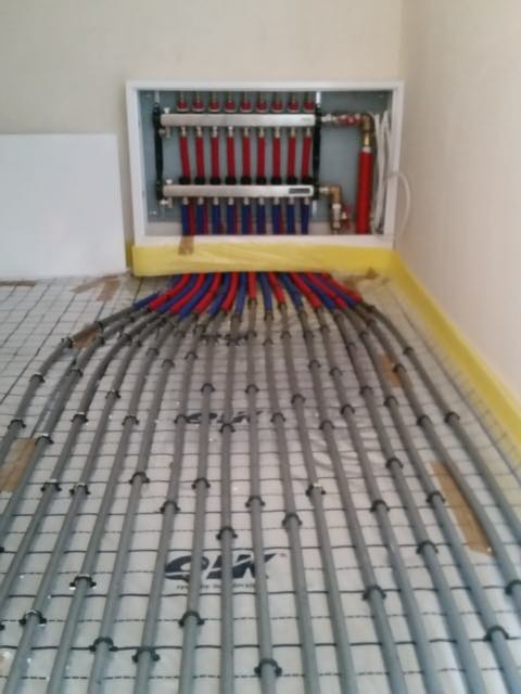 Rozdzielacz instalacji ogrzewania podłogowego z rurami grzewczymi w kolorze czerwonym i niebieskim, ułożonymi na izolacji podłogowej z siatką.