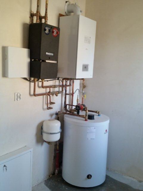 Zestaw urządzeń grzewczych: czarny moduł Buderus z czerwono-niebieskimi pokrętłami, biały kocioł grzewczy z kominem, biały zbiornik wyrównawczy i biały zasobnik ciepłej wody, połączone miedzianymi...