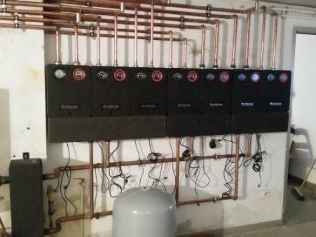 Rząd czarnych modułów grzewczych Buderus zamontowanych na ścianie, połączonych miedzianymi rurami i przewodami elektrycznymi, z widocznym zbiornikiem wyrównawczym i fragmentem ściany...