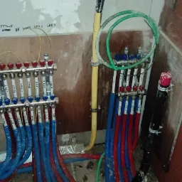 Rozdzielacze ogrzewania podłogowego z rurami w kolorze niebieskim i czerwonym, zamontowane na ścianie z widocznymi napisami odręcznymi, w surowym stanie wykończenia.