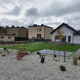 Nowoczesny ogród z szarą nawierzchnią żwirową, równo przystrzyżonym trawnikiem, młodymi drzewkami podpartymi palikami i tarasem z szarych płyt, na tle zabudowy mieszkalnej w pochmurny dzień.