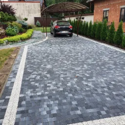 Ułożona kostka brukowa w odcieniach szarości z białym pasem oddzielającym, prowadząca do wiaty samochodowej z czarnym autem, otoczona zielenią z elementami dekoracyjnymi.