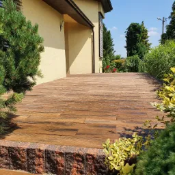 Rustykalny taras z desek kompozytowych, obrzeże z ceglastych bloczków, otoczony zielenią i niskimi iglakami, widok na elewację budynku.