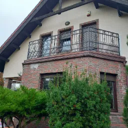 Kuta balustrada balkonowa z dekoracyjnymi elementami na tle domu z ceglaną elewacją i zielenią. Ujęcie z dołu podkreśla detale balustrady i architekturę budynku.