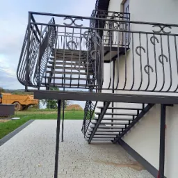 Zewnętrzne schody metalowe z balustradą, zdobione motywem podkowy, prowadzące na balkon. W tle widoczny krajobraz i maszyna budowlana.