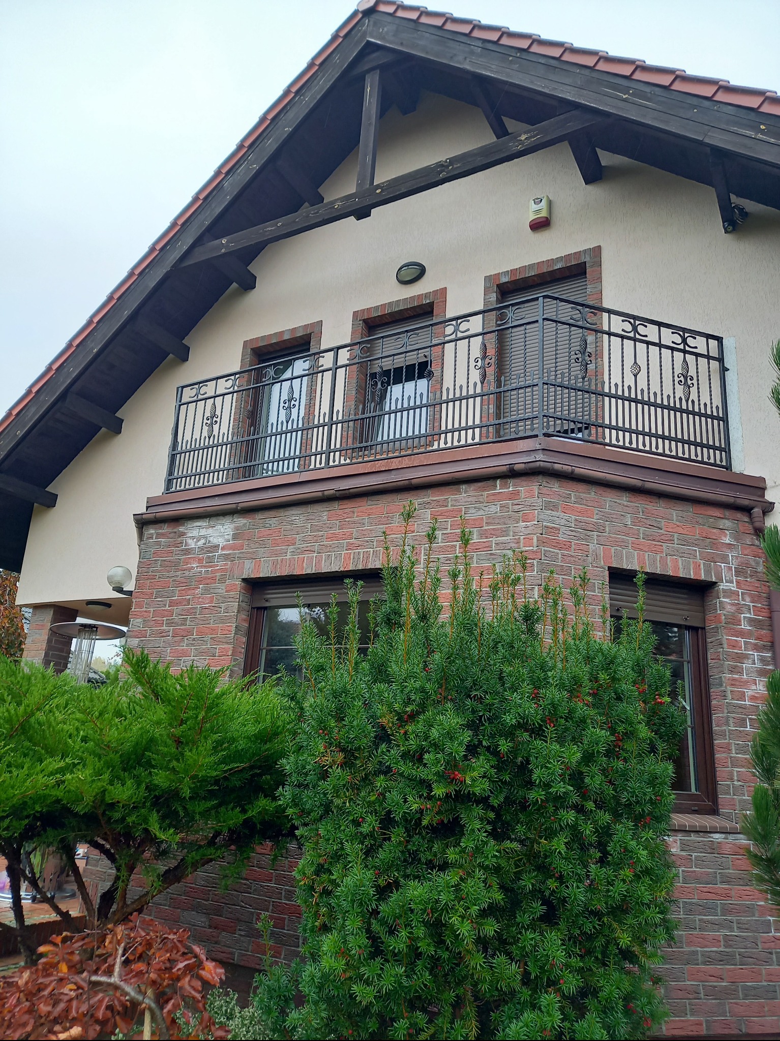 Kuta balustrada balkonowa z dekoracyjnymi elementami na tle domu z ceglaną elewacją i zielenią. Ujęcie z dołu podkreśla detale balustrady i architekturę budynku.