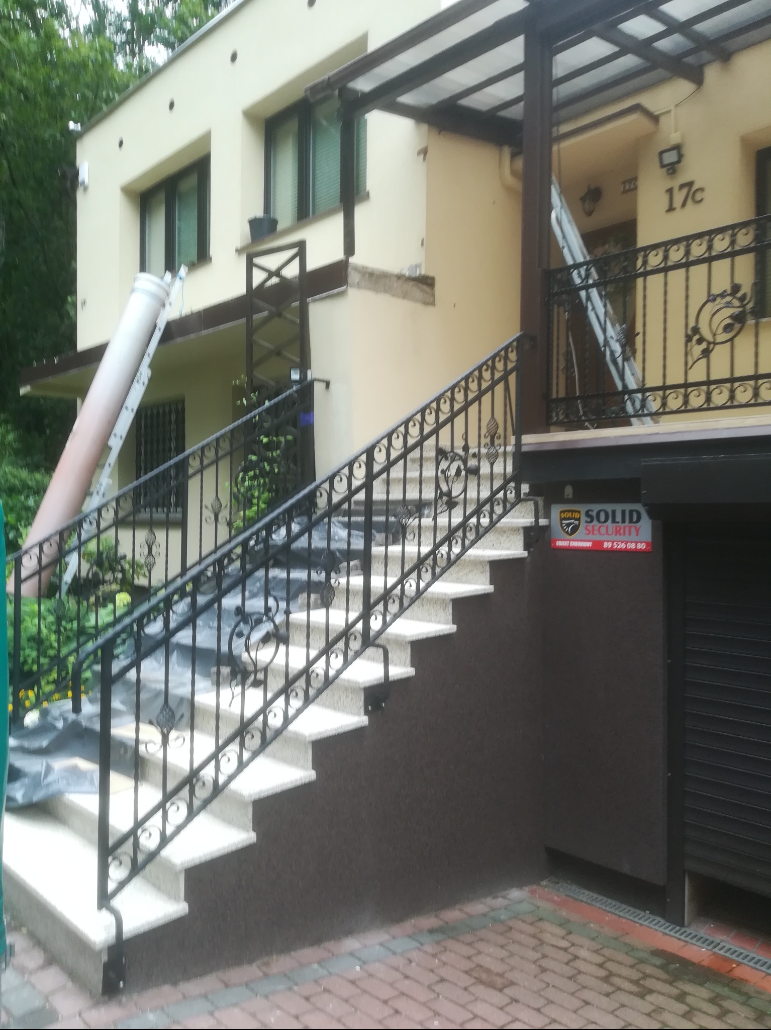 Zewnętrzne schody z kutą balustradą w kolorze czarnym, prowadzące do wejścia budynku. Widoczna drabina oparta o elewację i elementy rusztowania.