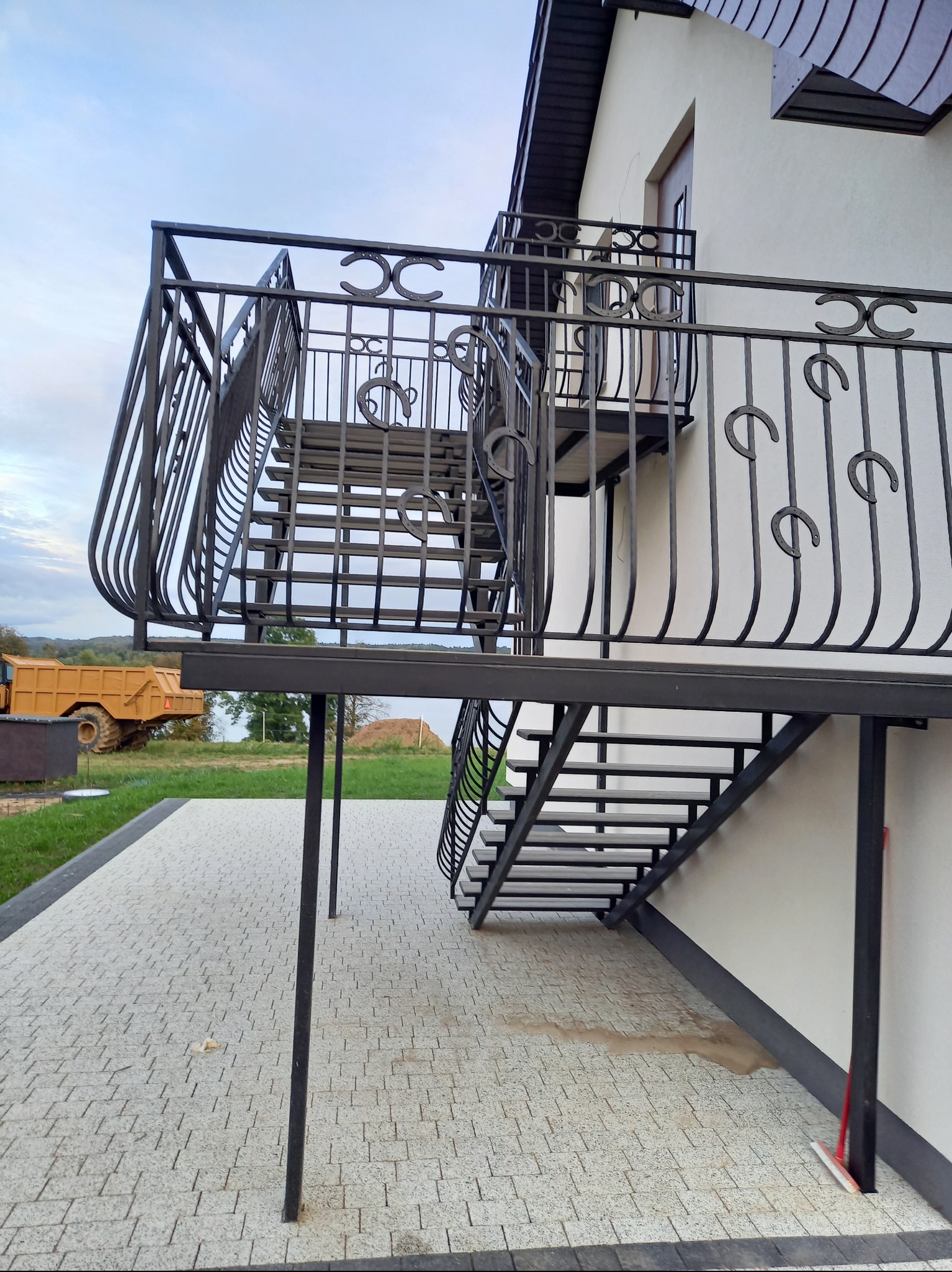 Zewnętrzne schody metalowe z balustradą, zdobione motywem podkowy, prowadzące na balkon. W tle widoczny krajobraz i maszyna budowlana.
