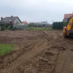 Żółta koparko-ładowarka JCB na polu w Pułtusku, przygotowanym pod zasiew, widoczne ślady opon na mokrej ziemi, w tle zabudowa jednorodzinna.