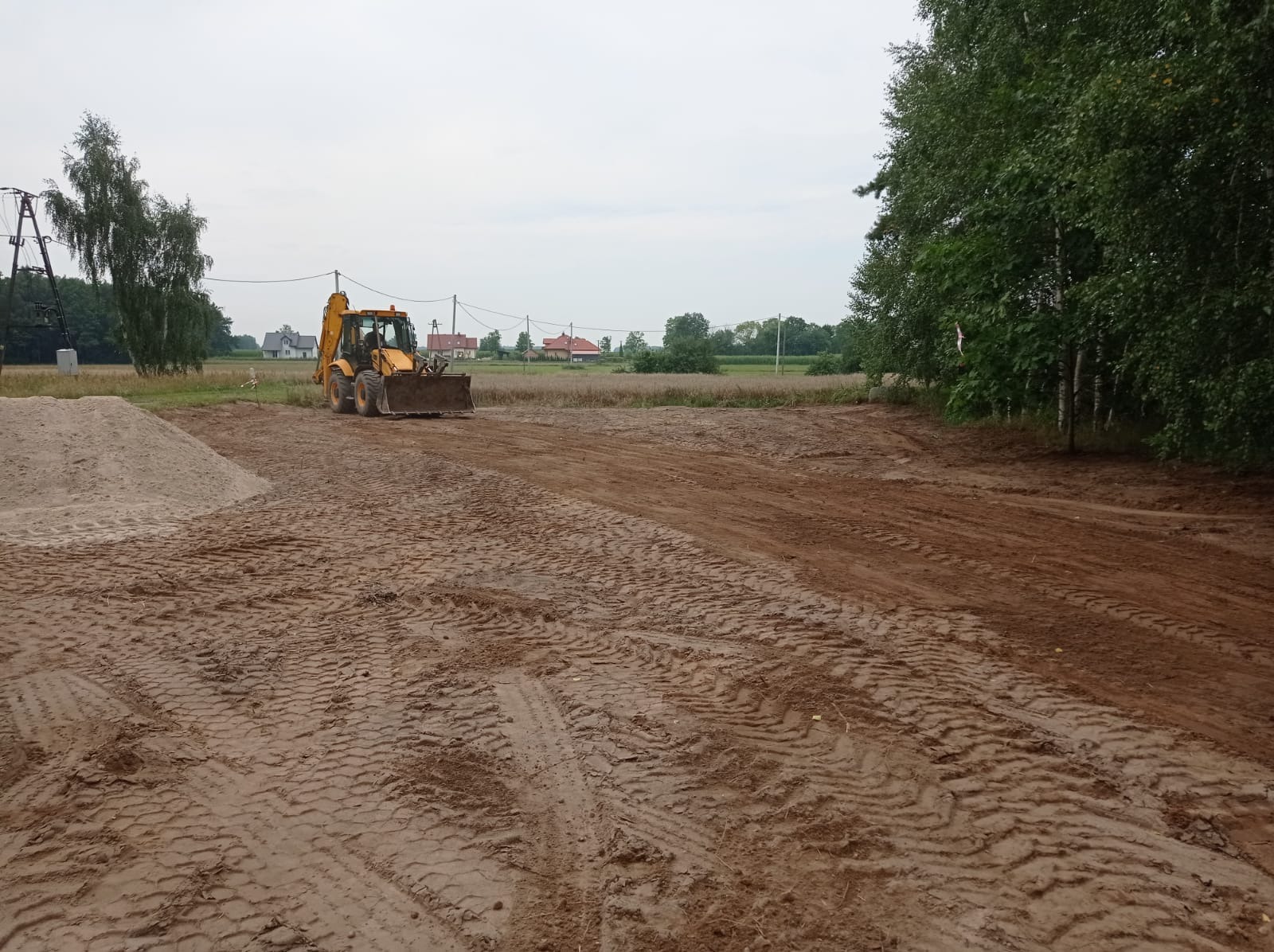 Żółta ładowarka kołowa wyrównuje teren pod budowę, widoczne ślady opon na ubitej ziemi i pryzma piasku w tle.