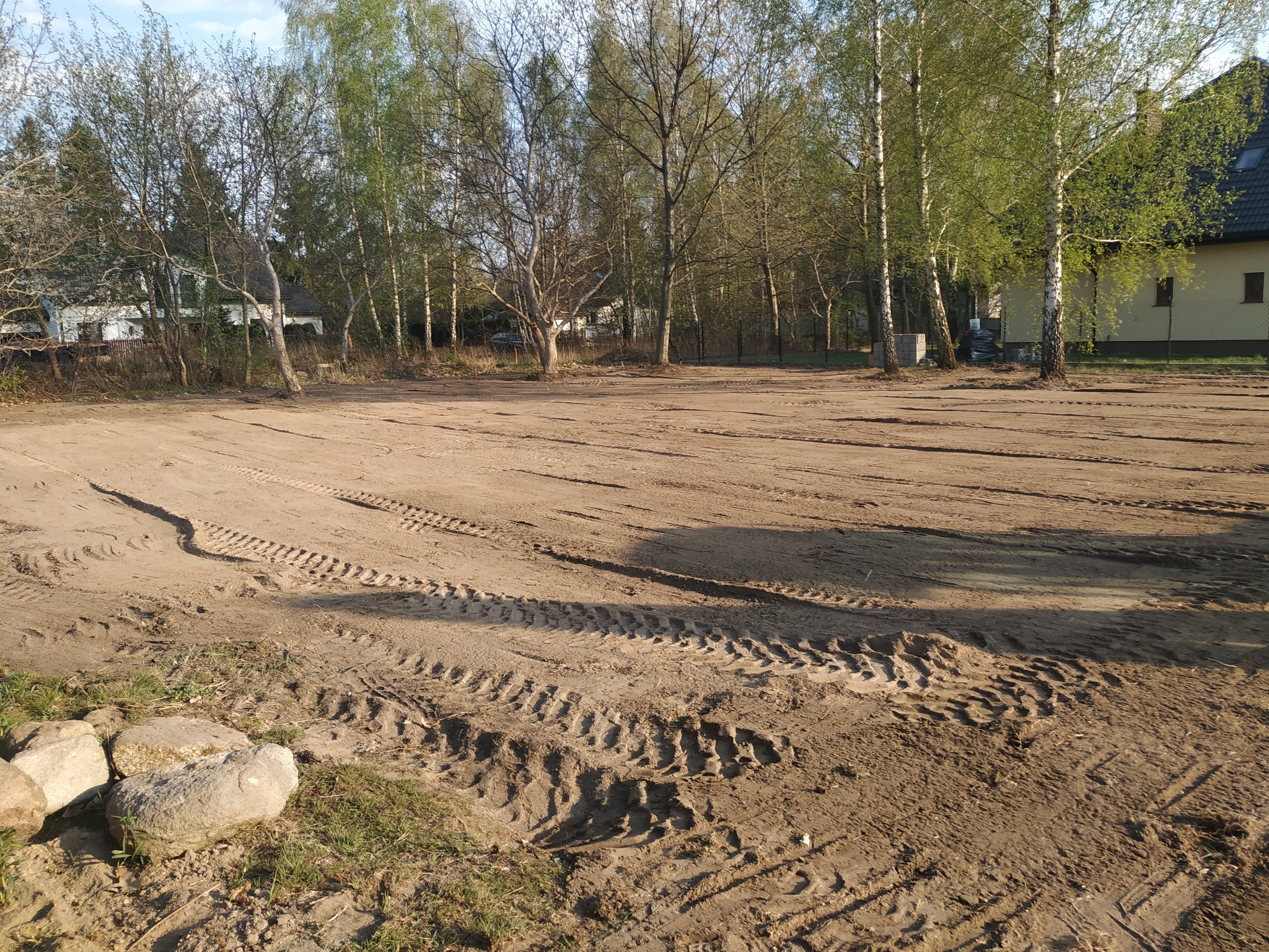 Wyrównany teren pod budowę z widocznymi śladami opon, w tle drzewa i budynek mieszkalny.