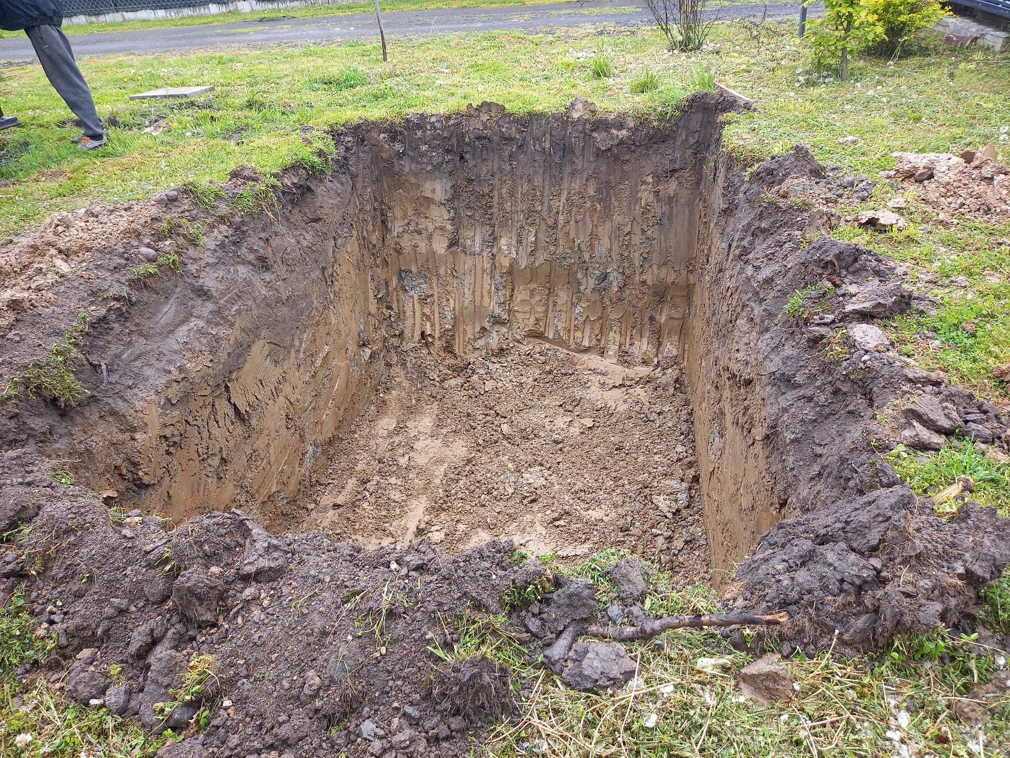 Prostokątny wykop w ziemi z pionowymi ścianami, otoczony darnią i ziemią, widoczne ślady narzędzi na ścianach wykopu.