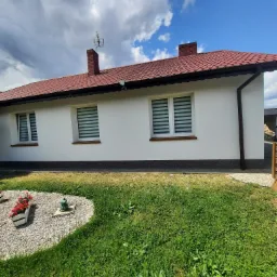 Biały dom z czerwoną dachówką i trzema oknami z żaluzjami, otoczony zielonym trawnikiem z dekoracyjnym klombem z kwiatami i kamykami.