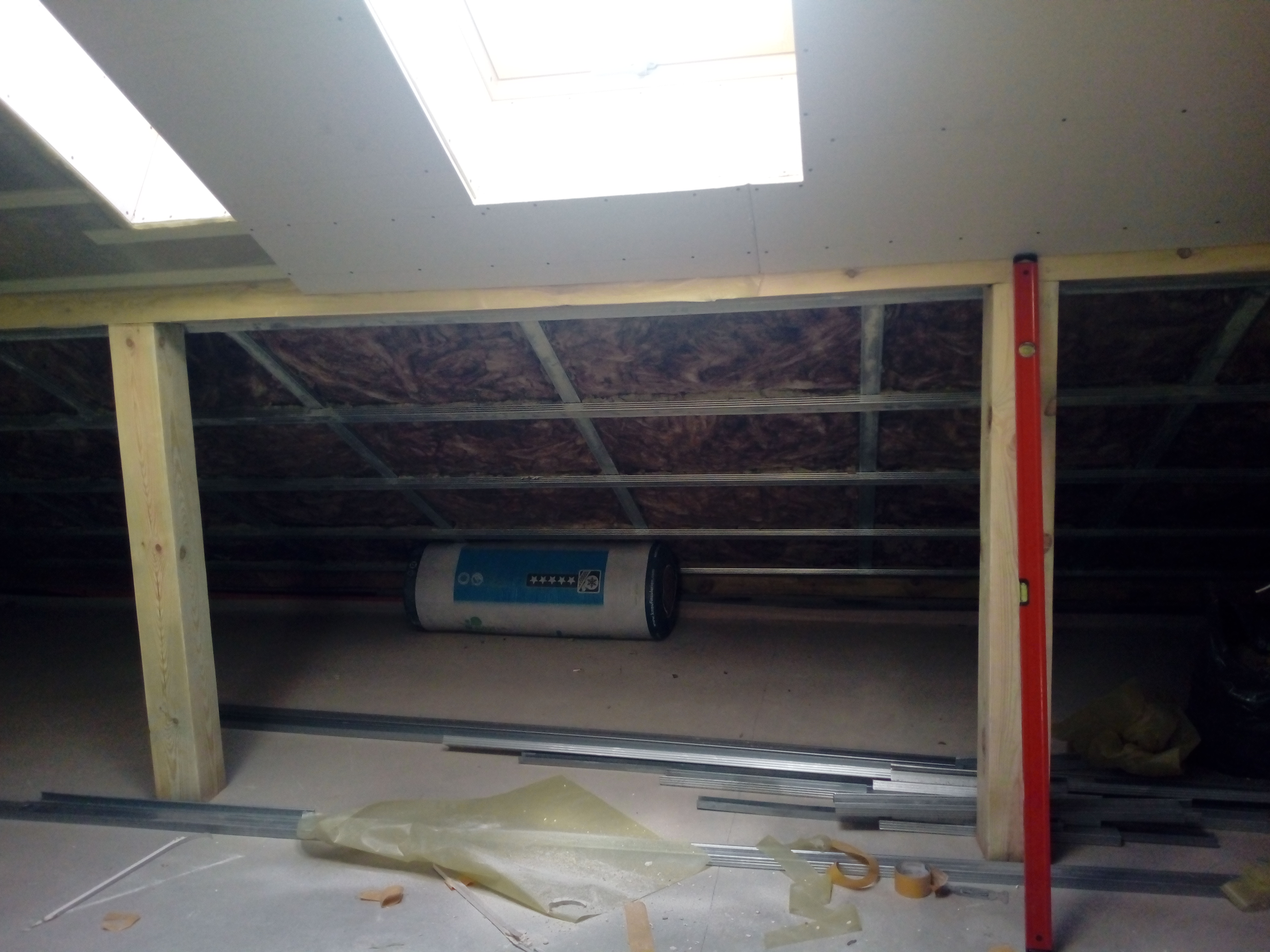 Poddasze w trakcie adaptacji: widoczne ocieplenie wełną mineralną, konstrukcja z profili stalowych, częściowo zamontowane płyty gipsowo-kartonowe z oknami dachowymi oraz poziomica przy pionowym...