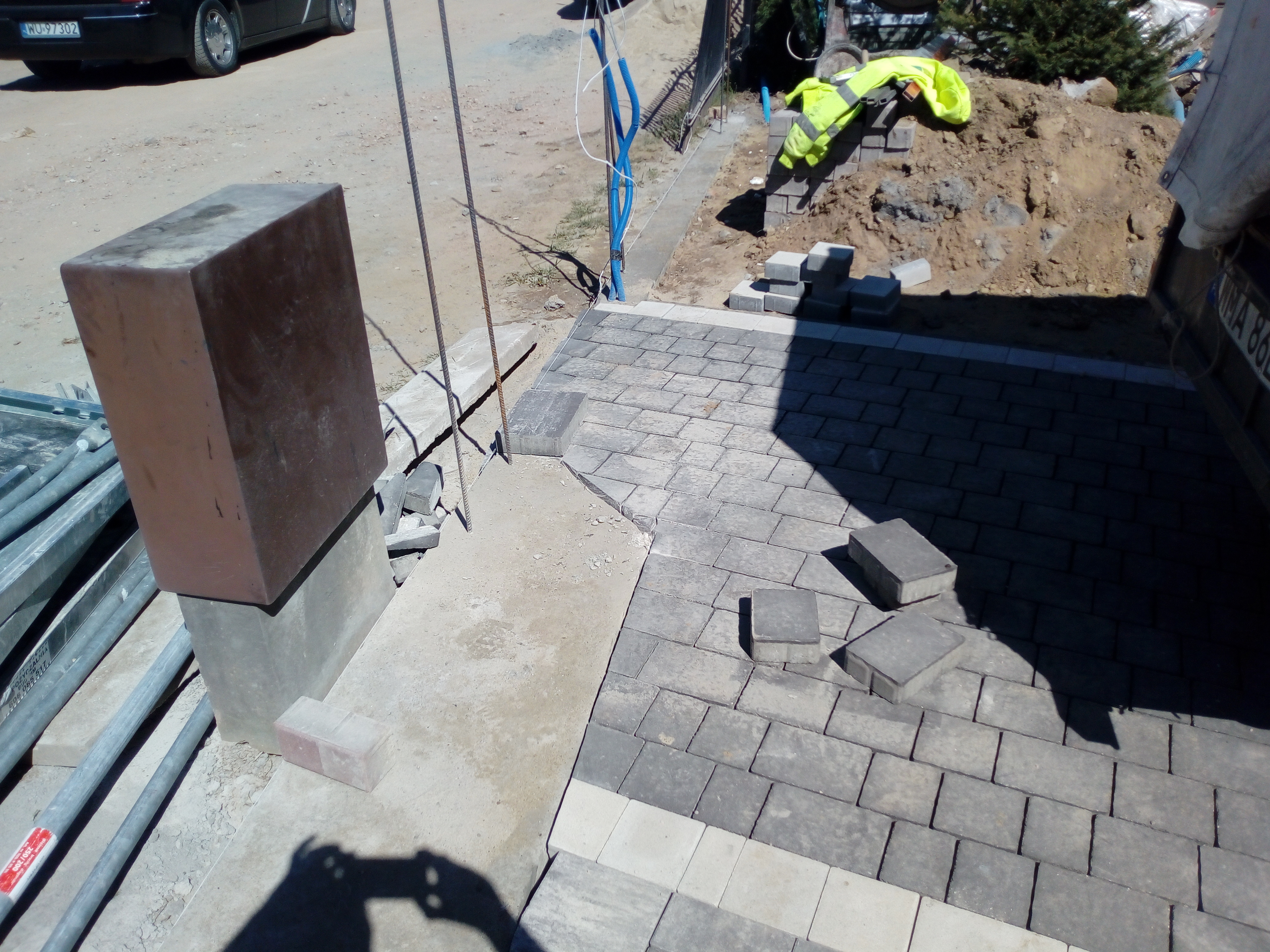 Trwa układanie szarej kostki brukowej z jasną obwódką na podjeździe, widoczne przygotowane kostki, słupek ogrodzeniowy, oraz odblaskowa kurtka na pryzmie ziemi w tle.