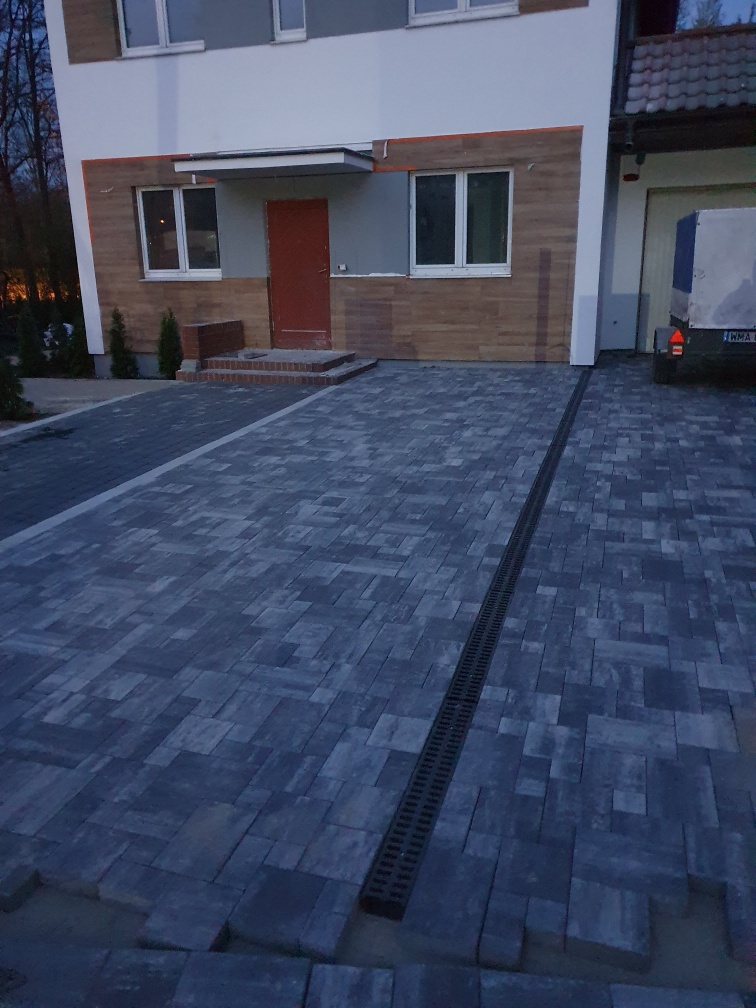 Ułożona kostka brukowa w odcieniach szarości z odpływem liniowym na posesji przed budynkiem z elewacją imitującą drewno.