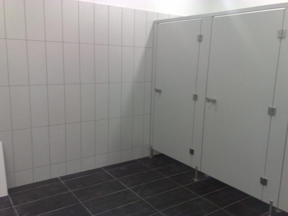 Publiczna toaleta z kabinami, wykończona białymi płytkami ściennymi i ciemną, kwadratową podłogą. Widoczne metalowe okucia i nóżki kabin.